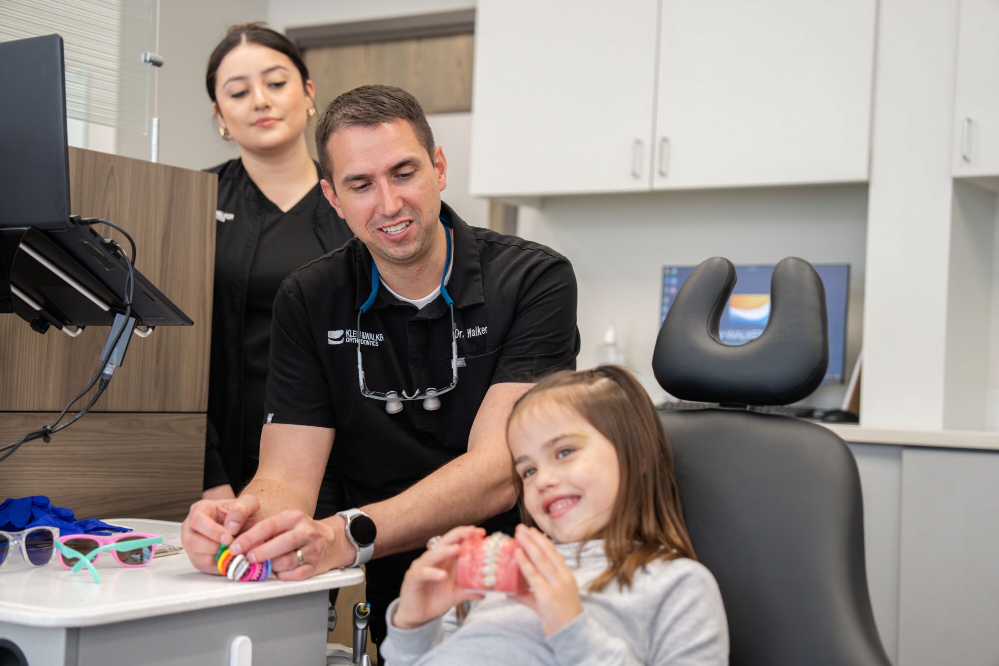 Child smiling while holding a model of braces, with Dr. Walker and an assistant in a modern orthodontic office, showcasing personalized orthodontic care in West Olathe.