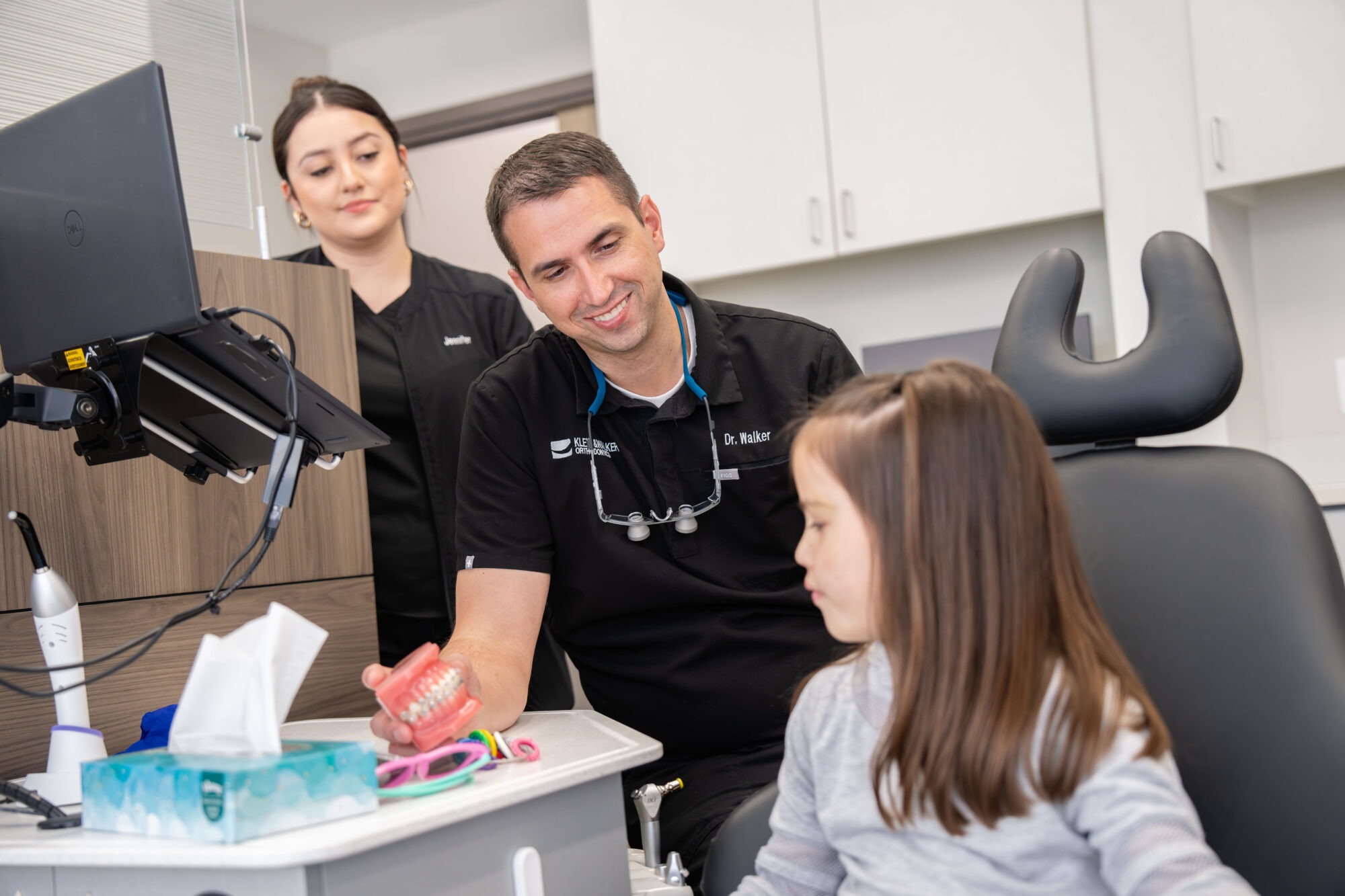 Child consulting with Dr. Walker at Klein Walker Orthodontics, examining orthodontic model, with dental assistant in background, emphasizing personalized orthodontic care for children.